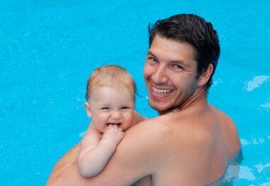 Ein Vater mit einem Baby auf dem Arm steht mit dem Rücken zur Kamera und dreht seinen Kopf über die Schulter, damit er in diese schauen kann. Beide lächeln.