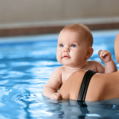 Zu sehen ist eine Mutter, die ihr Baby im Wasser auf dem Arm hält. Lediglich die Schulter der Mutter ist von hinten zu sehen. Das Baby schaut in Richtung Kamera.